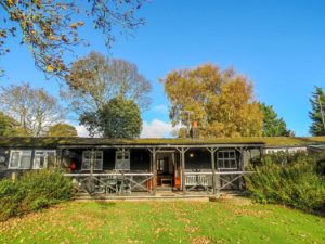 7 Uplands, two bedroom cottage in Thorpeness - black timber bungalow with covered veranda and lawn under autumn trees