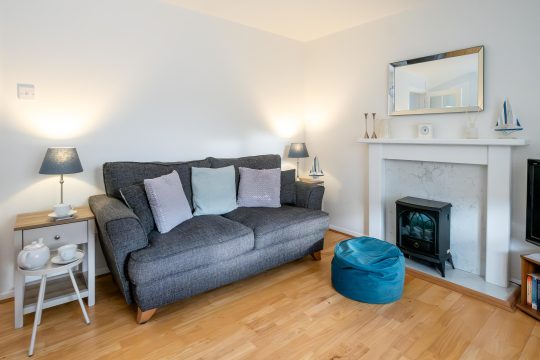 The Britten, one bedroom apartment in Aldeburgh - living room with grey sofa, electric stove in a white fireplace and side tables