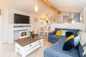 Beach Lodge, two bedroom lodge in Sizewell - open-plan living room with blue corner sofa, electric stove and kitchen beyond
