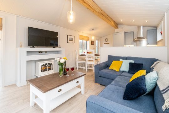 Beach Lodge, two bedroom lodge in Sizewell - open-plan living room with blue corner sofa, electric stove and kitchen beyond